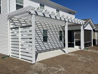 White pergola and privacy wall attached to a house with a concrete patio; clear blue sky.