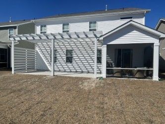 White pergola attached to a white house with a covered patio, set in a yard.