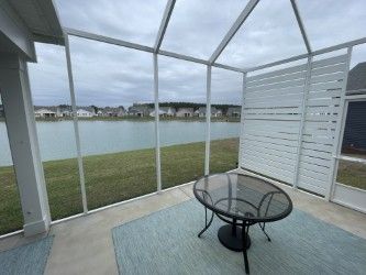 Screened-in porch with lake view, featuring a glass-top table, blue rug, and white horizontal slat wall. Overcast sky.
