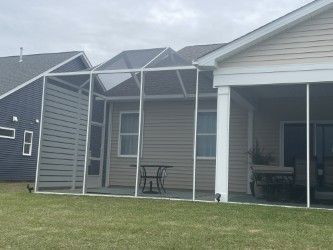 Screened-in porch attached to a beige house, with a table and chairs inside. Green grass in the foreground.
