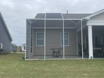 Screened-in patio with white frame. Beige house backdrop with table & chairs inside. Cloudy sky overhead, green lawn.