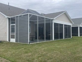 Screened porch attached to a light-colored house. Gray siding, white trim, and a peaked roof are visible.