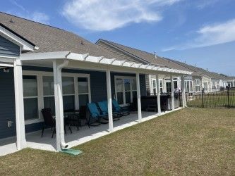 White pergola covers a patio attached to a blue house with chairs and a table.
