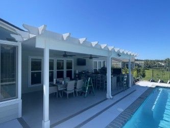 White pergola over a patio with dining table and outdoor kitchen, beside a pool.