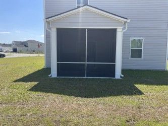 Screened-in porch attached to a light gray house. The porch has a white trim and dark screens. Green grass surrounds it.