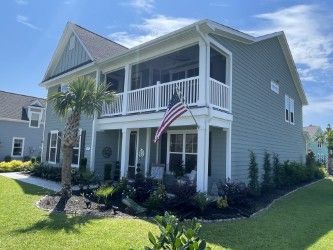 Two-story blue house with porch, white railings, American flag, palm tree, and green lawn.