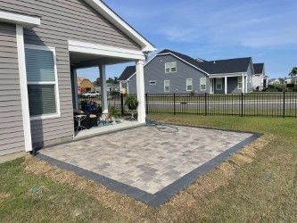 Paver patio outside a gray house with a black border, surrounded by grass and a black fence.