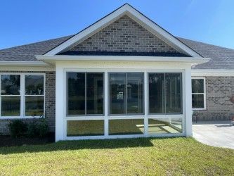 Sunroom addition to a brick house with large windows, white trim, and a gabled roof, set in a grassy yard.