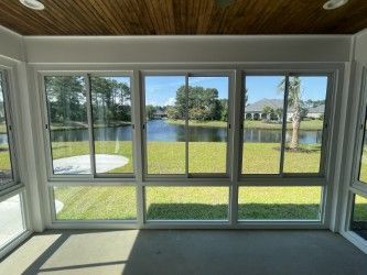 Sunroom with large windows overlooking a lake and green lawn. White frames, gray floor, brown ceiling.