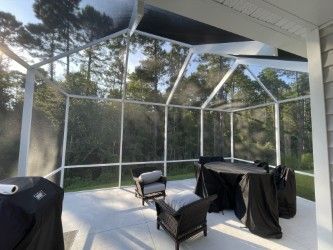 Screened-in patio with chairs and covered table, white frame, concrete floor, overlooking trees.