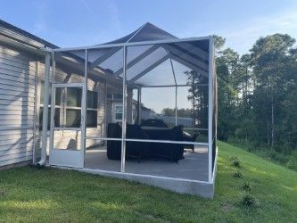 Screened-in porch attached to a house. White frame, gray concrete floor, dark furniture, green lawn and trees in background.
