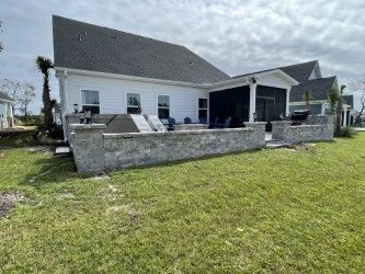 Backyard patio with gray brick wall, lounge chairs, and a white house with black roof, under a cloudy sky.