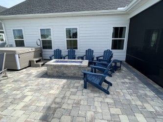 Patio with fire pit, blue Adirondack chairs, and hot tub near a white house.