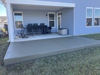 Gray concrete patio extends from a blue house with windows, covered by an overhang. Dark outdoor furniture sits on the patio.