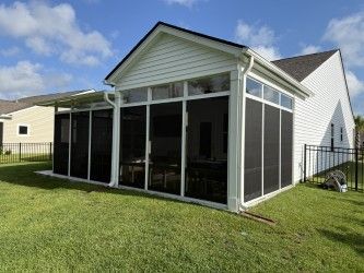 A screened-in porch with white frames is attached to a white house with a black roof, set on a grassy lawn under a partly cloudy sky.