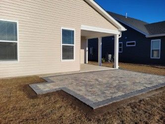 Exterior view of a house with a concrete patio. The patio is brick-like in pattern and bordered by dark pavers.