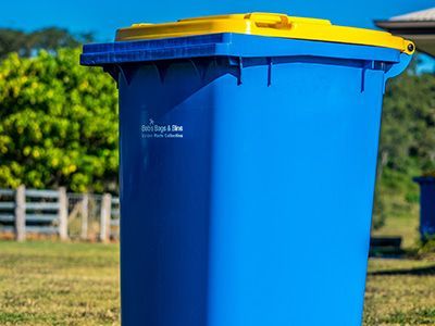 Blue and yellow recycling bin in a grassy yard with a fence and trees in the background.— Bobs Bags & Bins in Walkerston, QLD