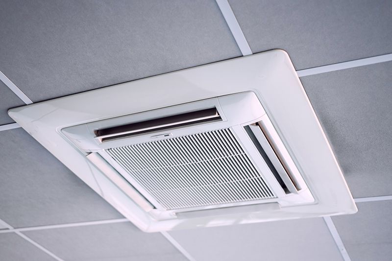 Ceiling-mounted air conditioner in a grid ceiling, white with vents and rectangular panel.