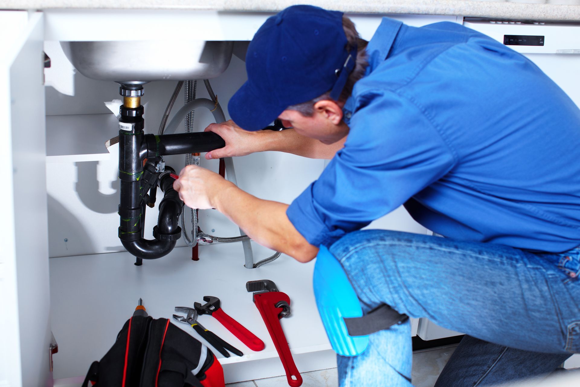 A plumber in a blue uniform fixing a kitchen sink with a wrench and tool bag nearby.