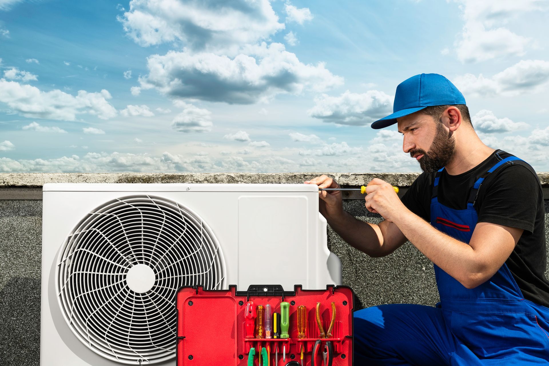 HVAC technician in blue overalls repairs an air conditioner unit on a rooftop with tool kit nearby.
