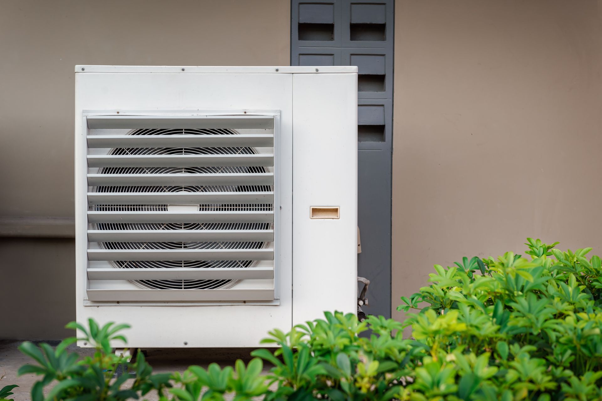 White HVAC unit with front vents, gray pillar, and green bushes against a tan wall.