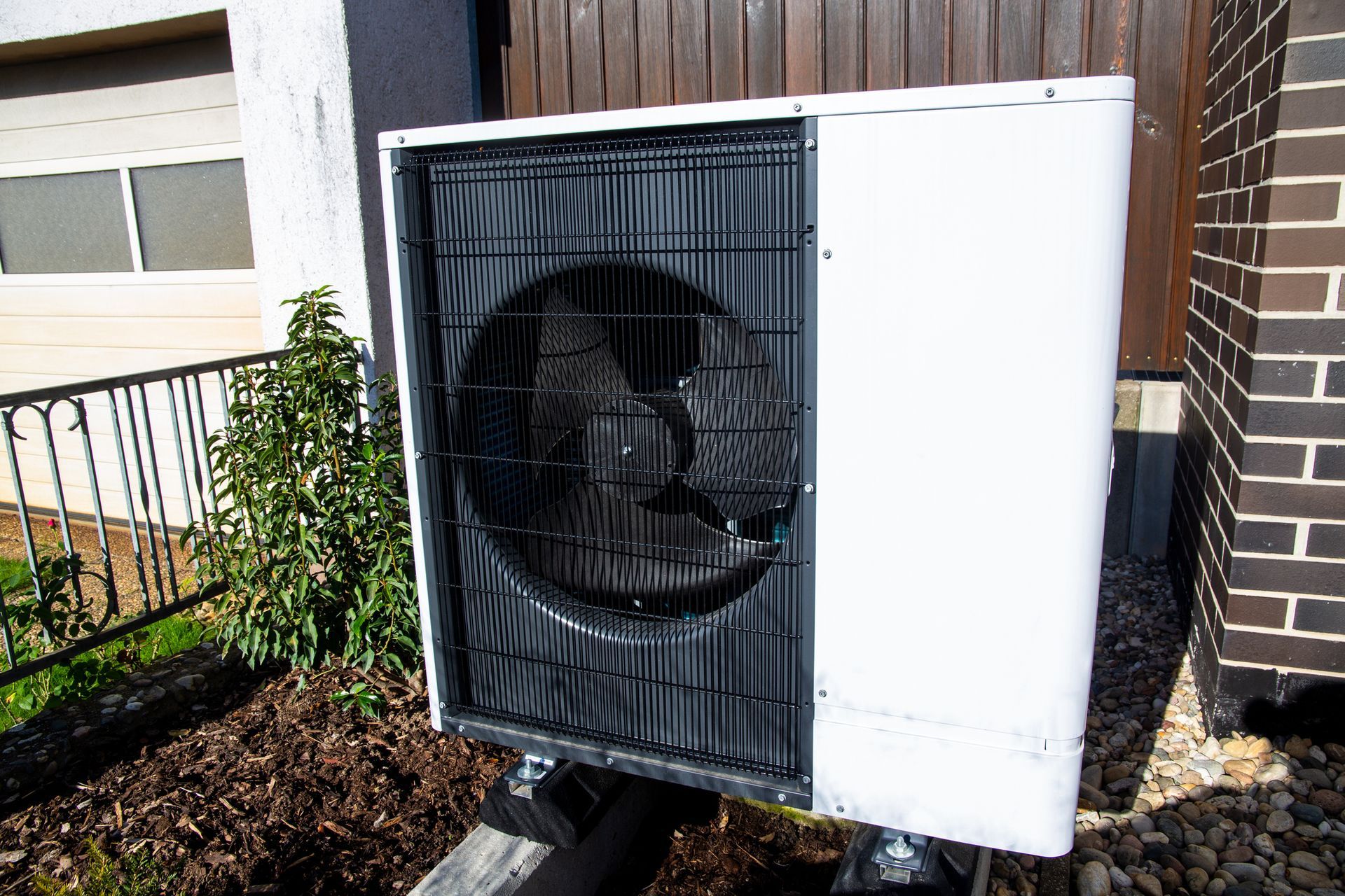 White heat pump unit outdoors, with a black fan behind a grid, near a brick wall and vegetation.