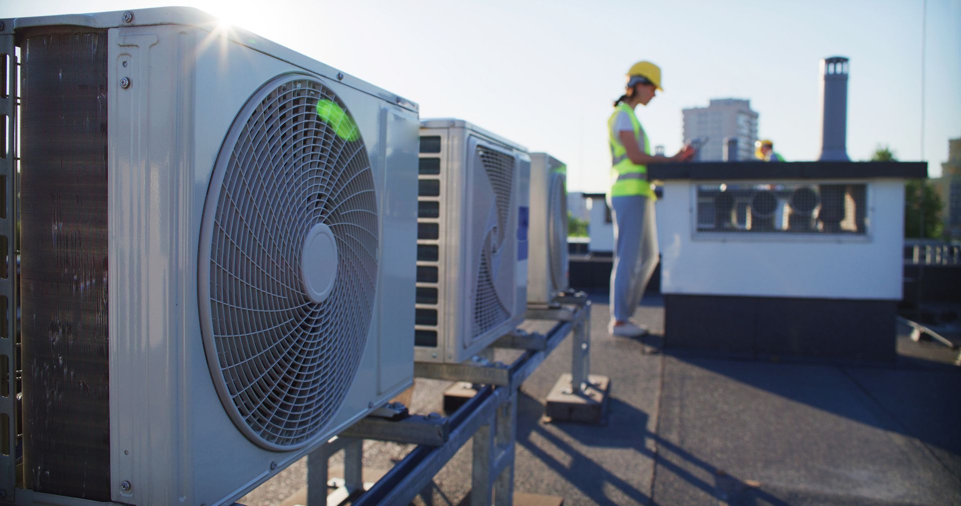 HVAC technician in a hard hat and safety vest on rooftop with air conditioning units. Sunny day.
