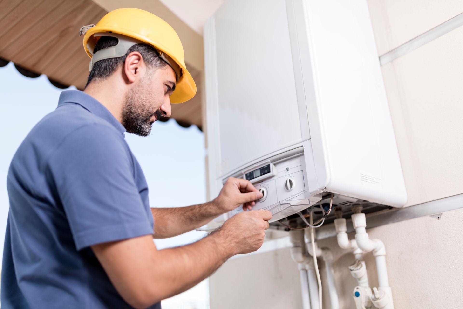 A repairman in a yellow hard hat adjusts a white wall-mounted boiler in a building.