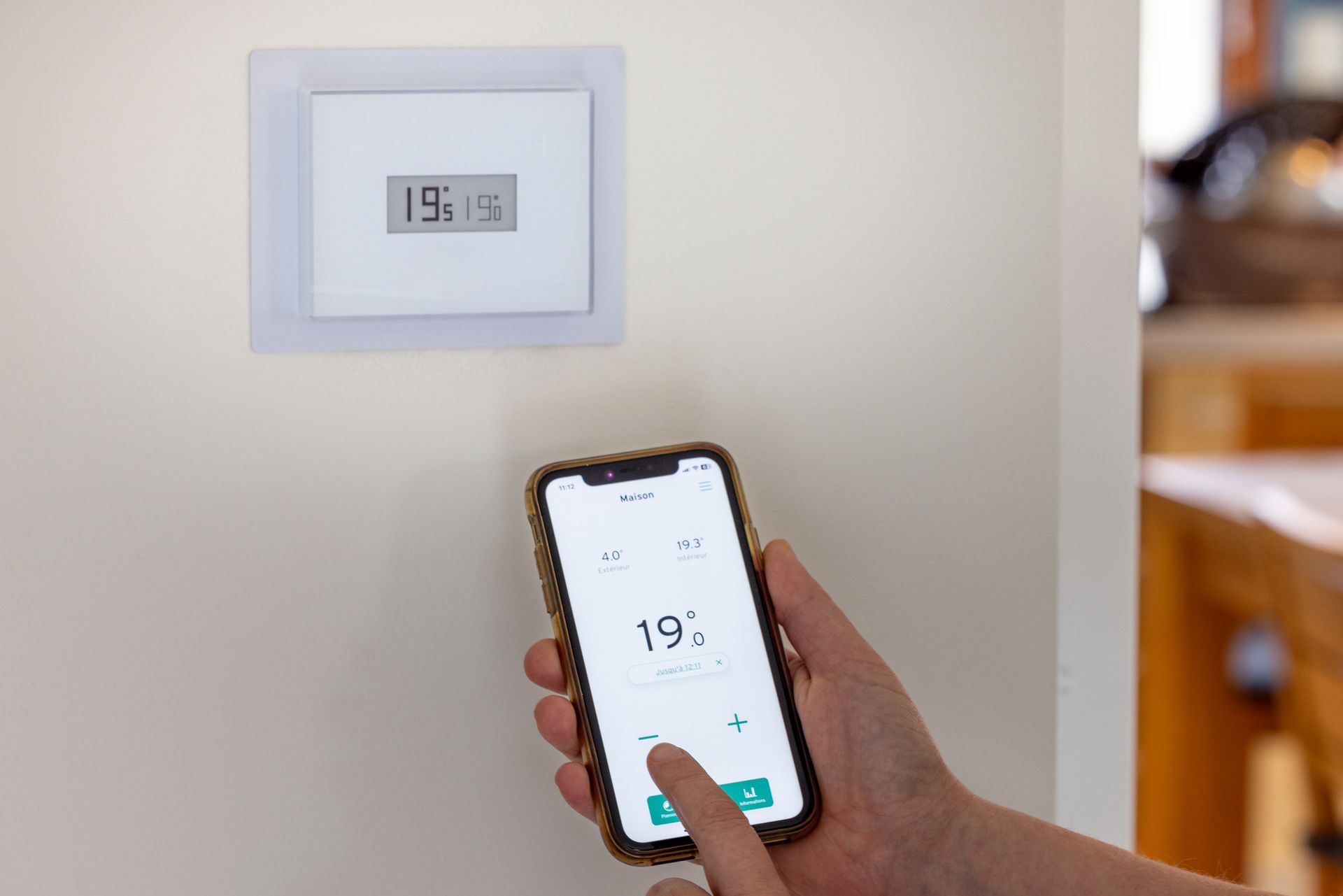 Hand adjusting a thermostat on a phone, with the same temperature displayed on a wall-mounted digital thermostat.
