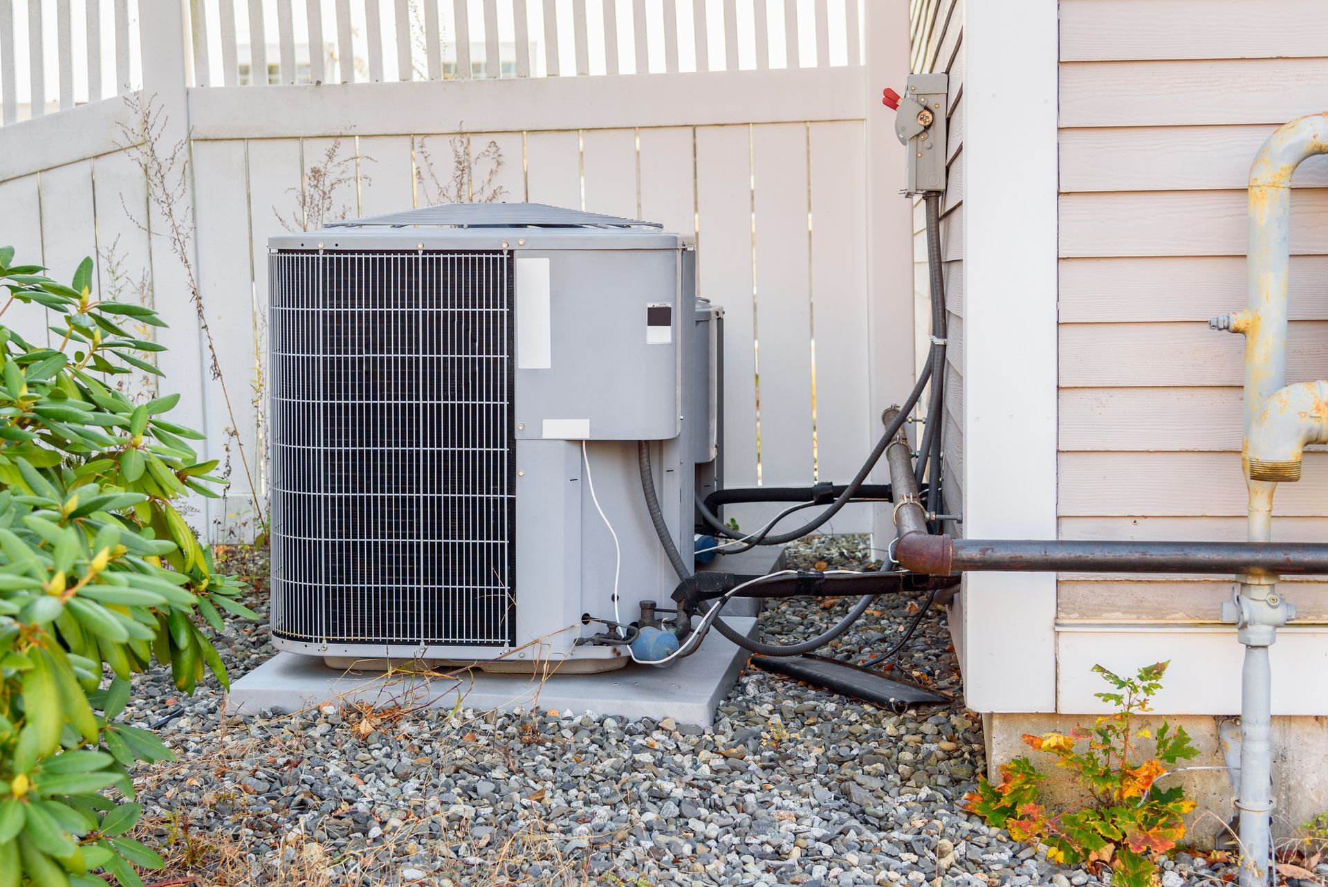 An outdoor air conditioning unit next to a white fence, nestled on a bed of gravel.