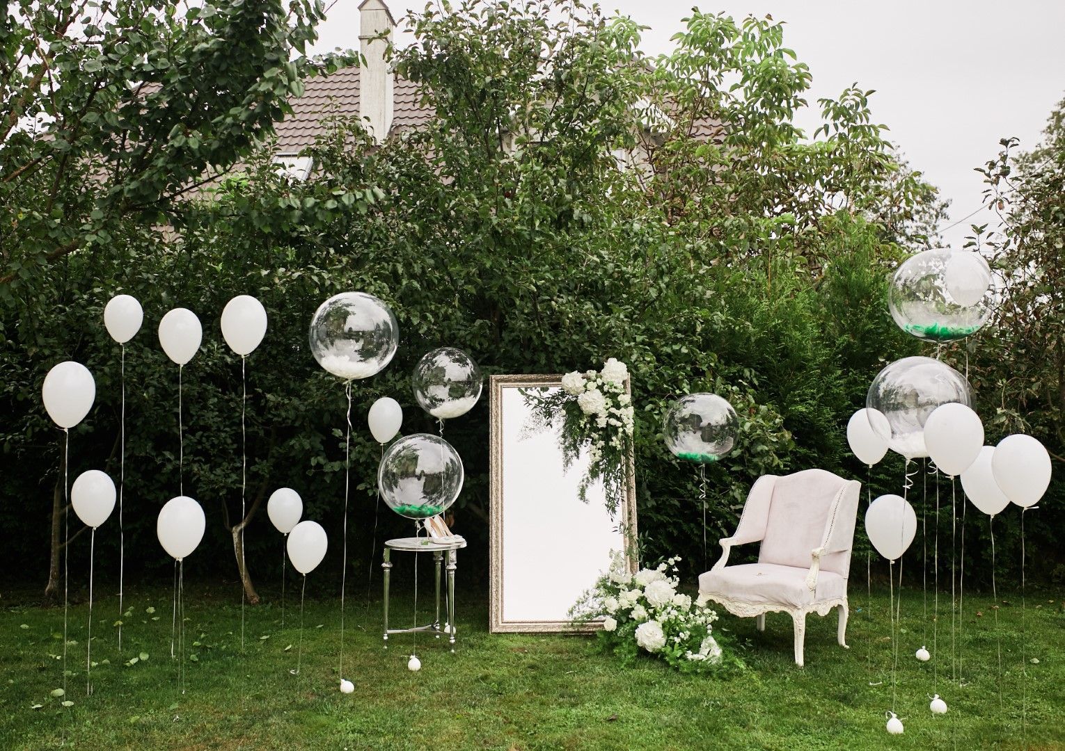 A chair and a mirror are surrounded by white balloons in a garden.