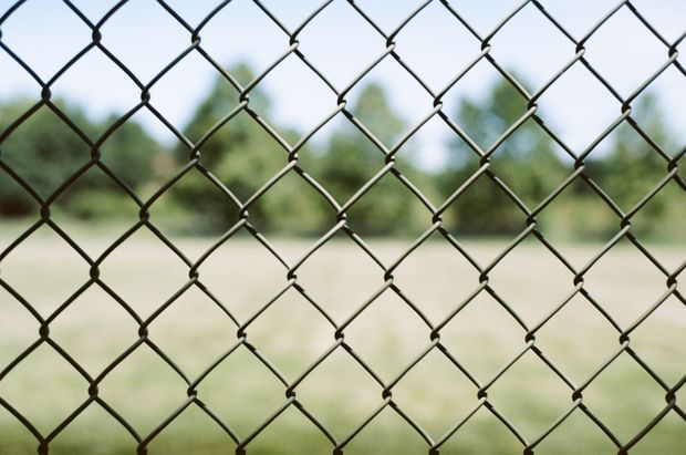 Construction site with chain-link fence, a tall building being demolished, and heavy machinery under a cloudy sky.