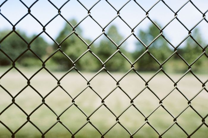 Construction site with chain-link fence, a tall building being demolished, and heavy machinery under a cloudy sky.