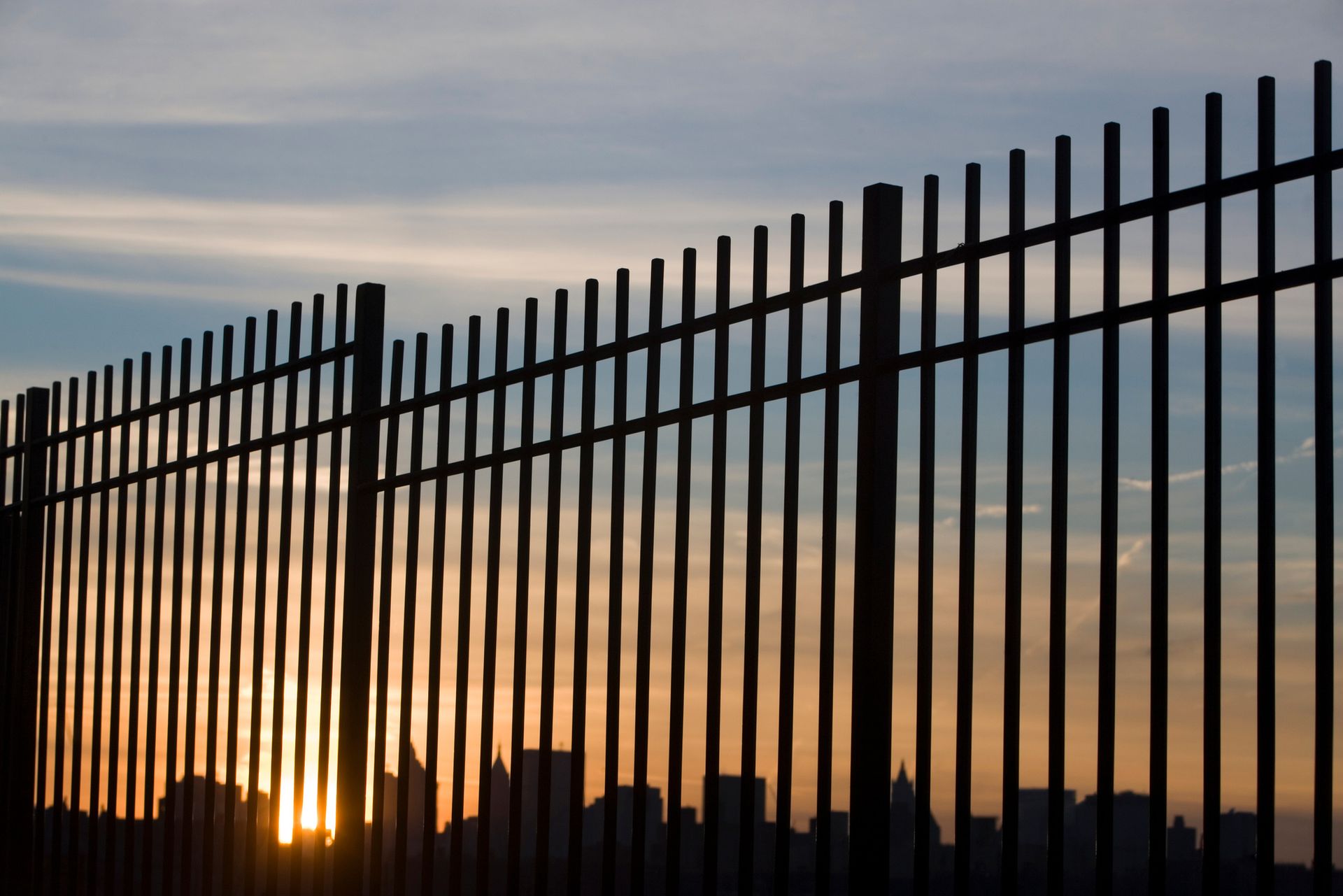 Silhouette of a metal fence against a sunset, with a cityscape visible at the bottom. Silhouette of a metal fence against a sunset, with a cityscape visible at the bottom.
