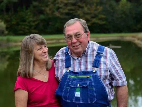 A couple stands near a pond. The man wears blue overalls, the woman a red shirt. Both smile. A couple stands near a pond. The man wears blue overalls, the woman a red shirt. Both smile.