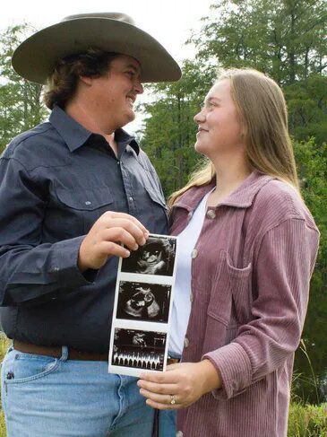Couple in field, holding ultrasound images. Man in cowboy hat, woman in pink shirt, both smiling.