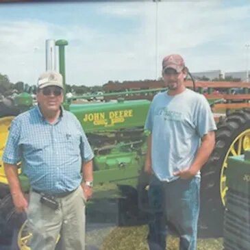 Two men stand by a green John Deere tractor. The older man wears a cap and checkered shirt, the younger a cap and t-shirt.