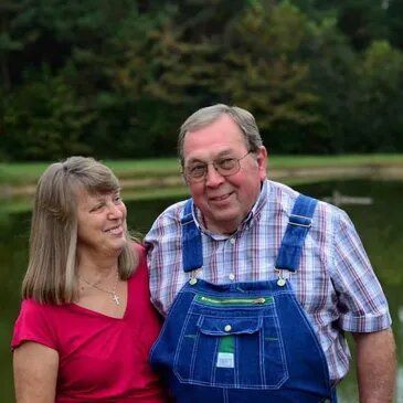 Man in blue overalls and woman in red shirt smiling by a pond.