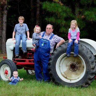 Family poses on and around a vintage tractor in a grassy field; blue overalls, smiles, and a rustic setting.
