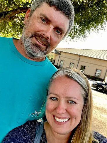 Man and woman smiling in selfie; man has grey beard, blue shirt; woman has headband.