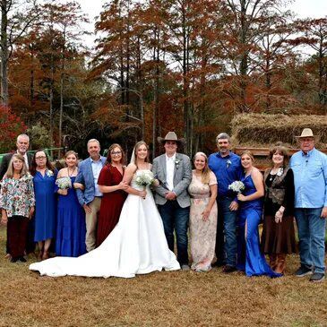 Wedding group posing outdoors; bride in white gown, groom in a hat, surrounded by family, fall foliage.