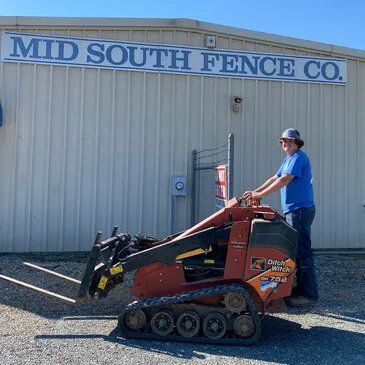 Man operating orange Ditch Witch mini skid steer in front of Mid South Fence Co. building.