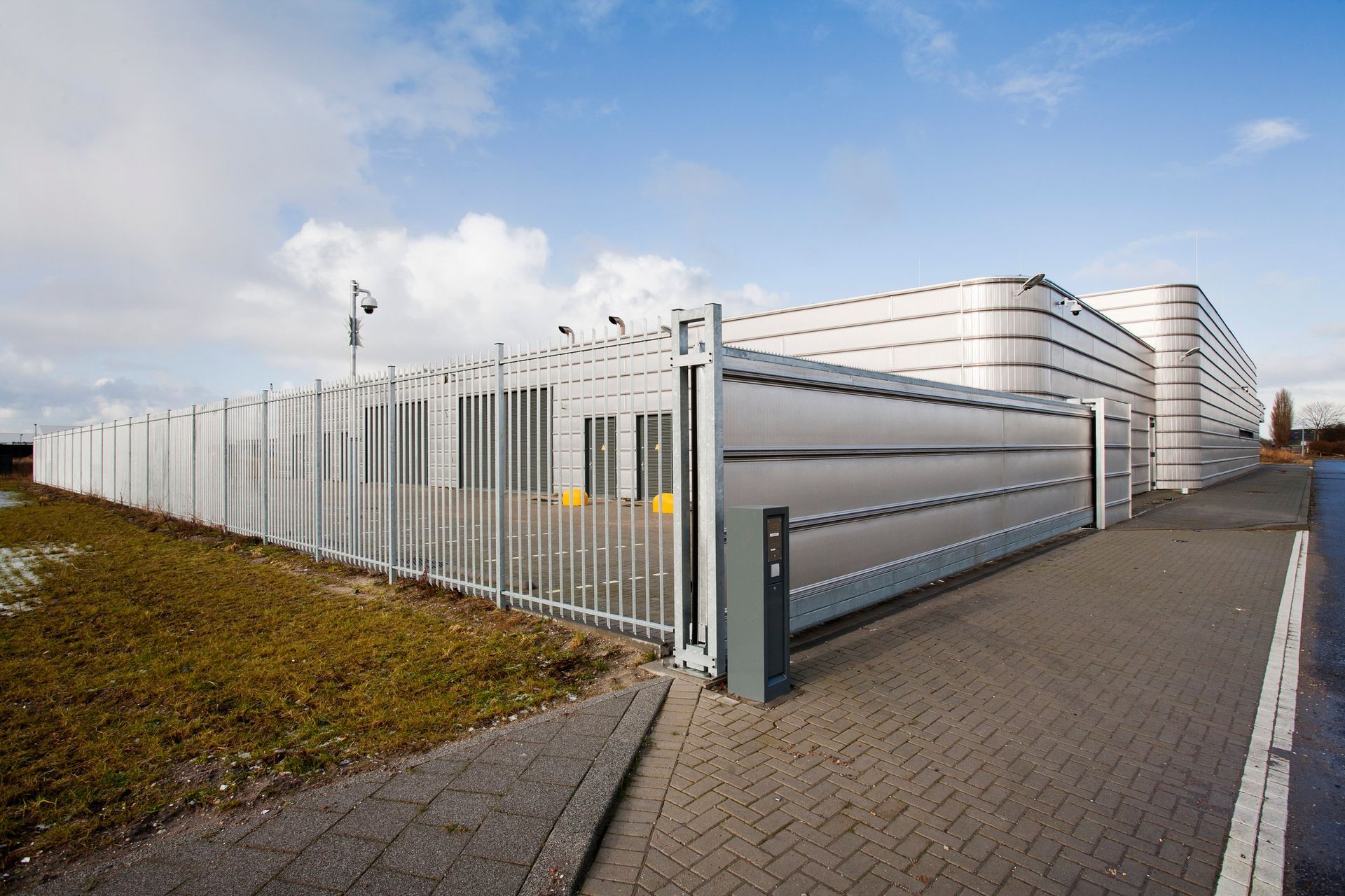 A modern, gray-walled building with a security fence under a cloudy blue sky.