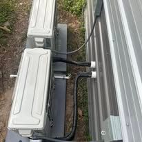 Two white air conditioning units mounted on a trailer next to a metal wall, connected by black tubes.