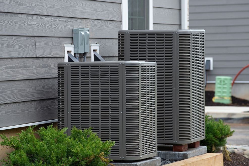 Two dark gray air conditioning units outside a gray house, surrounded by green bushes.