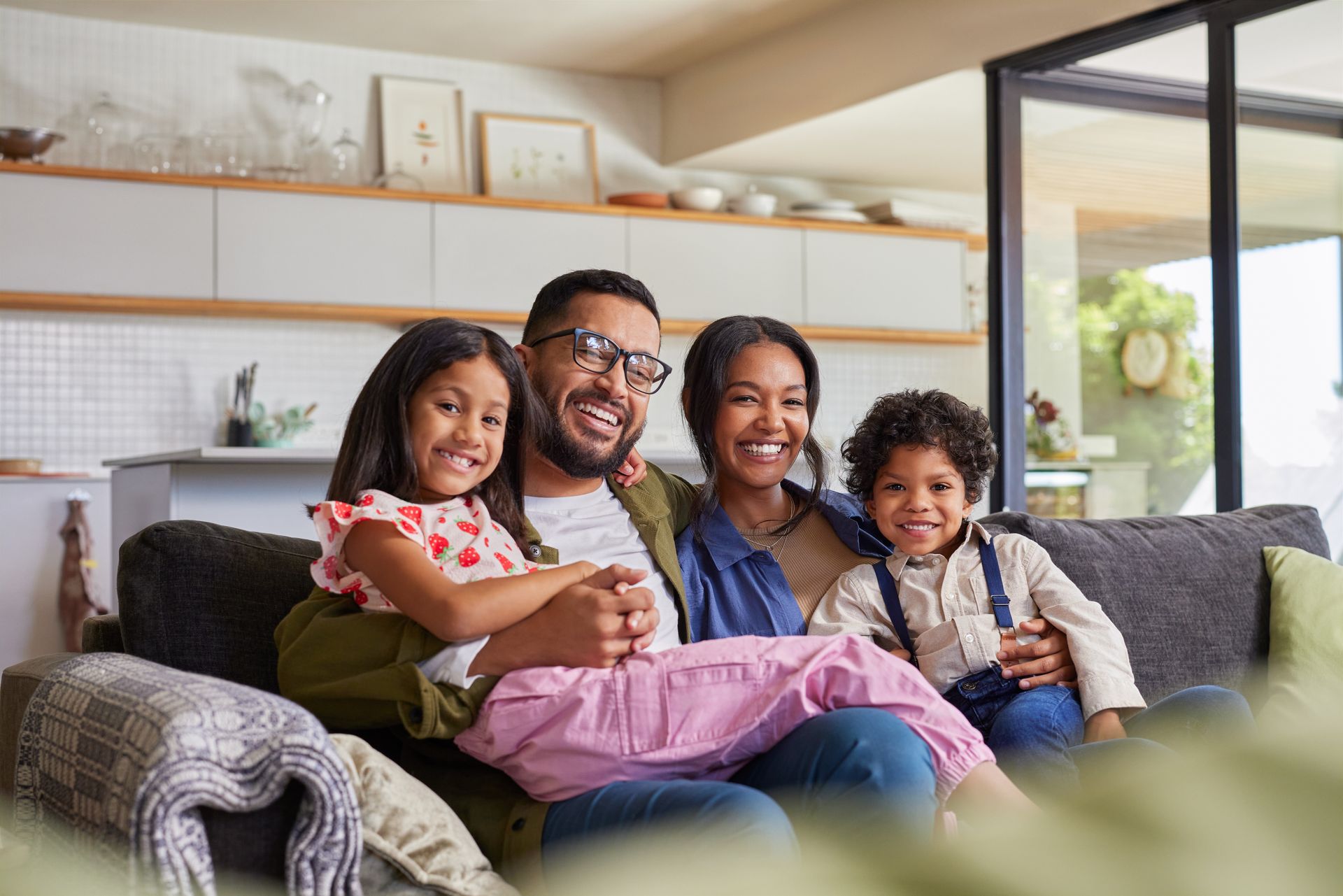 Family smiling on a couch together: a man and woman, two children. Modern living room setting.