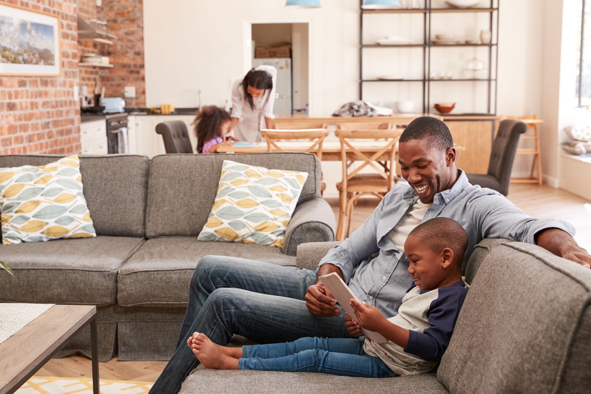 Father and son smile while looking at a tablet on a couch, mother and daughter in background in home.