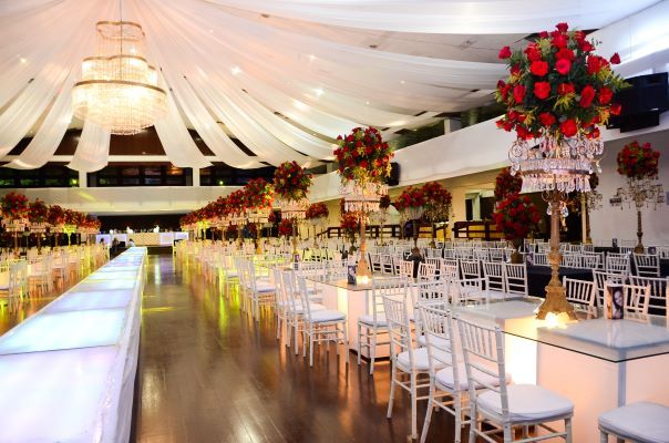 A large banquet hall with glass or white tables, and large flower arrangements