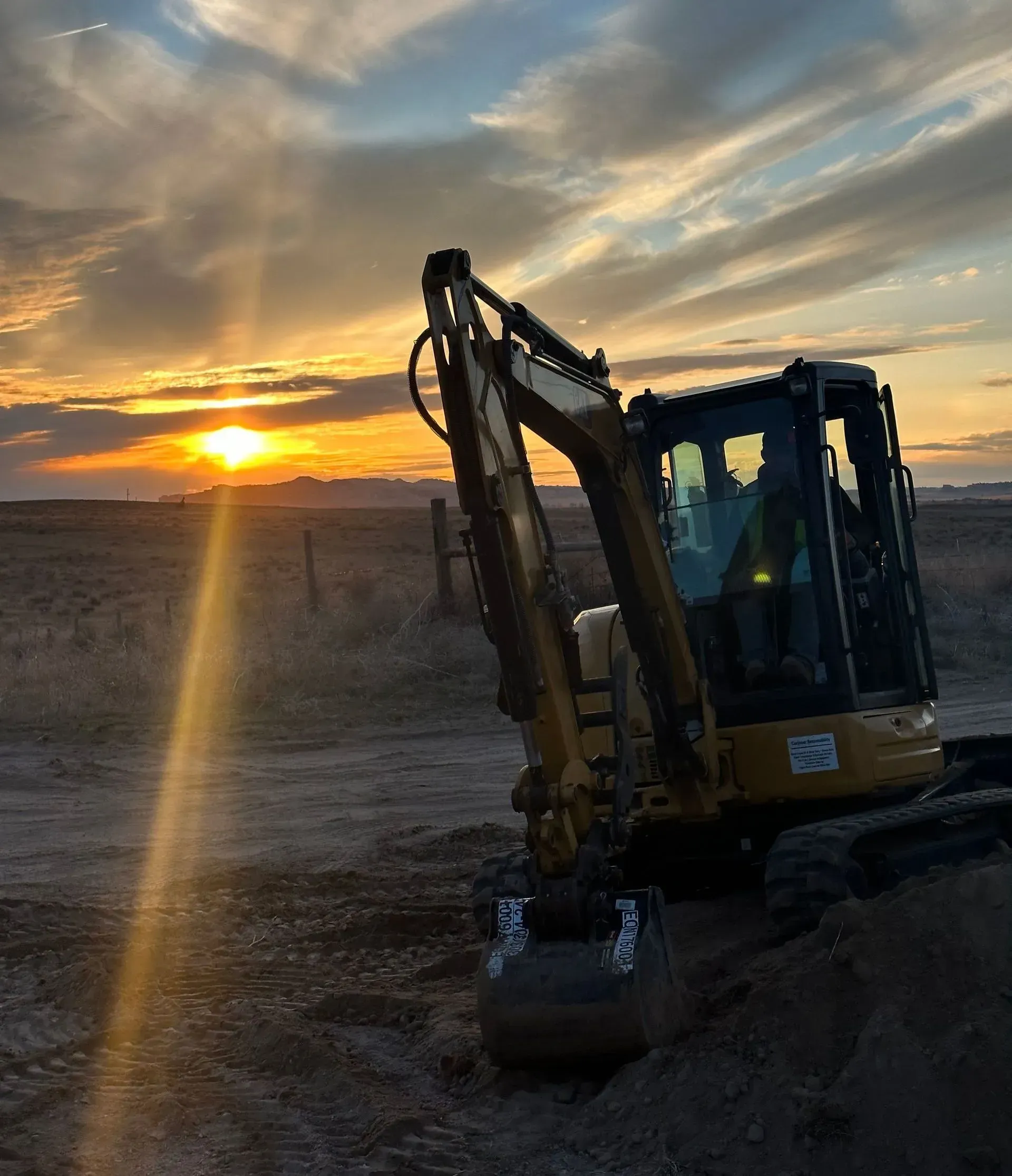 Yellow excavator at sunset, digging in a field, with a colorful sky.