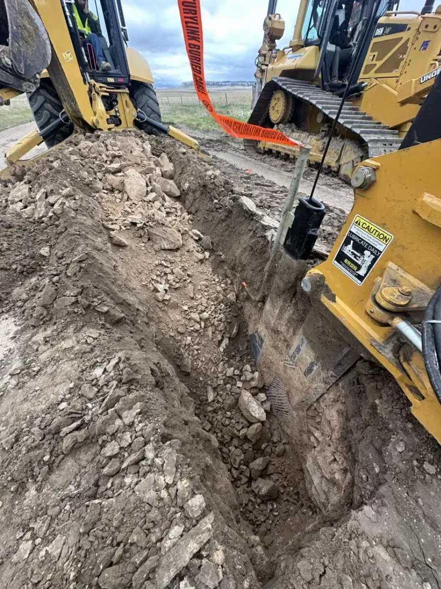 Yellow construction vehicles digging a trench in dirt, with orange safety tape overhead.